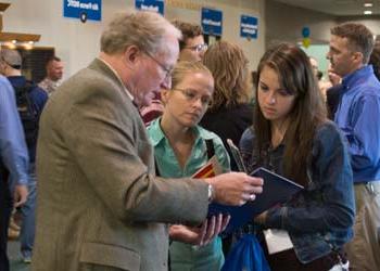 A young woman assists a girl with her homework at a small table in a room full of students.